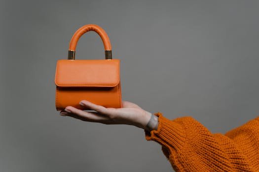 Stylish orange handbag held by a hand against a gray background. Perfect for modern fashion.